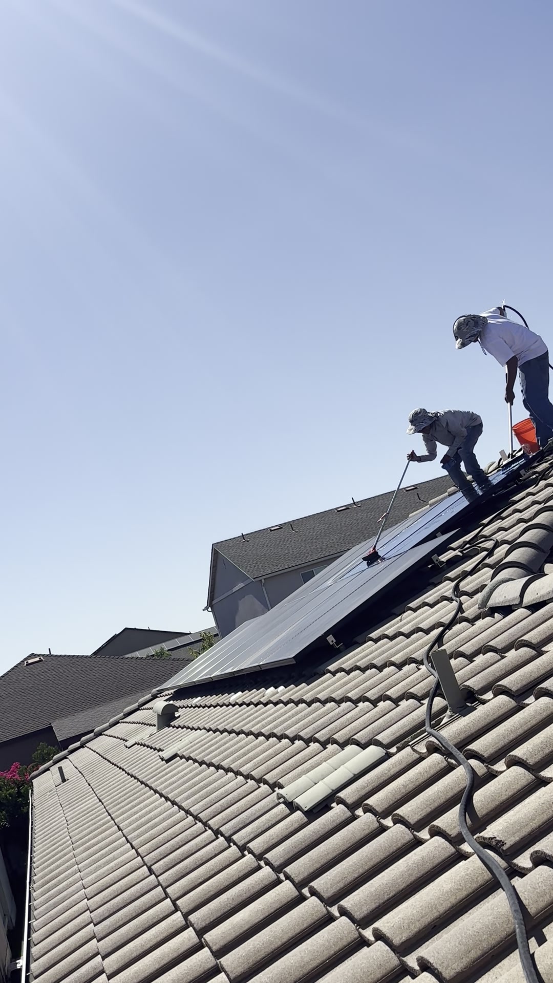 Technician reaching across a tile roof to soft-wash a residential solar array in action.