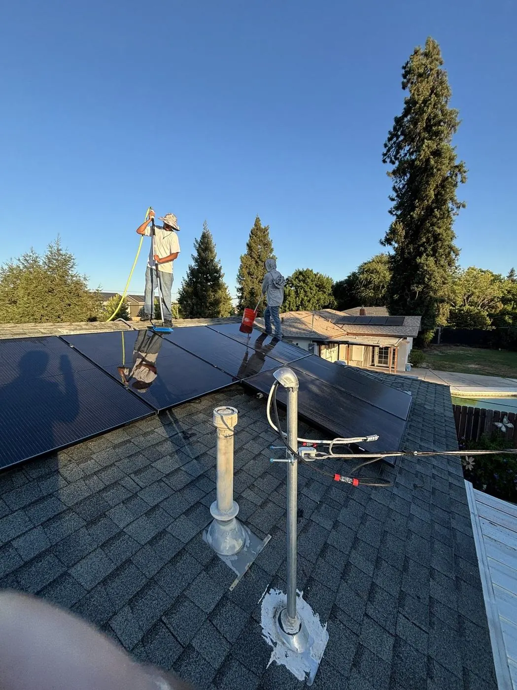 Technician cleaning rooftop solar panels with a water-fed brush pole under a clear blue sky.