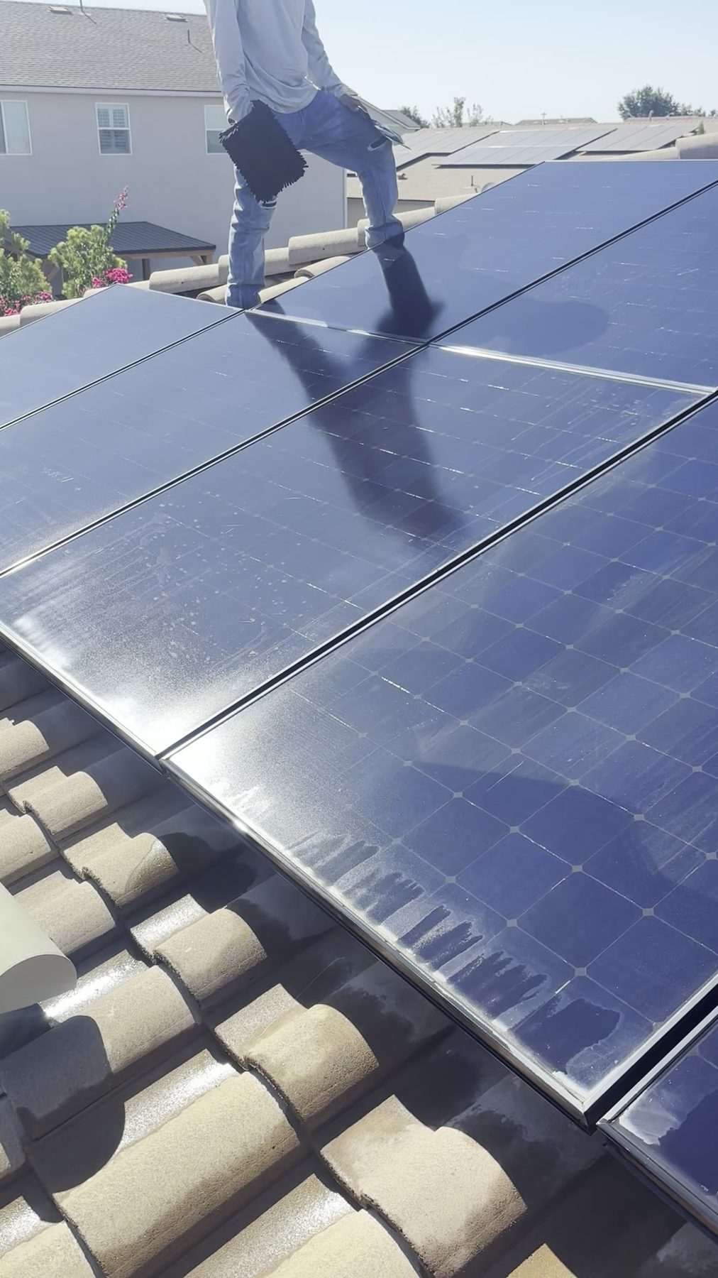 Technician standing on a roof cleaning solar panels above a Central Valley neighborhood.